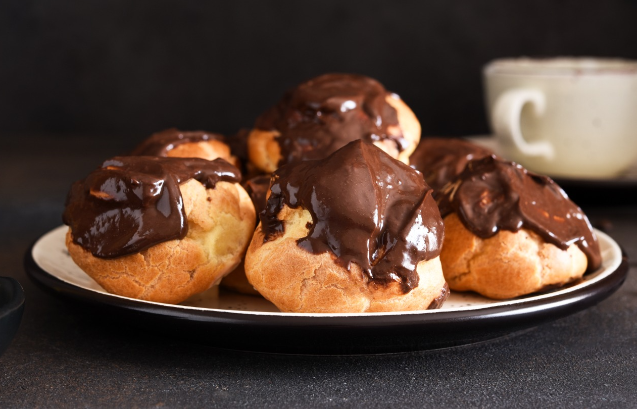 profiteroles-com-creme-de-baunilha-e-calda-de-chocolate-com-uma-xicara-de-cafe-na-mesa-da-cozinha.png